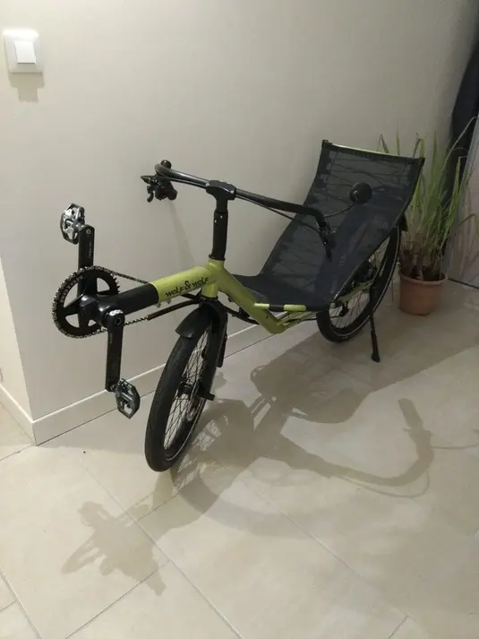 A lime green recumbent bicycle leaning against a white wall indoors on a light-colored tiled floor. The bicycle features a mesh seat back, black components, and the distinctive laid-back riding position of recumbent designs. A small potted plant is visible in the corner of what appears to be a home interior.