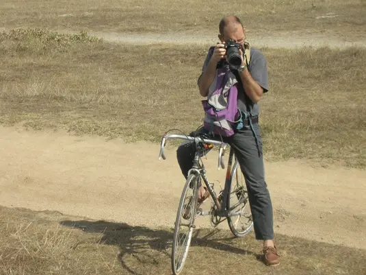 A person standing with a traditional upright bicycle on a dirt path in a dry, grassy area. The person is holding a camera up to their face, taking a photo. They're wearing a gray shirt, dark pants, and brown shoes. A purple bag or item is visible on the bicycle.
