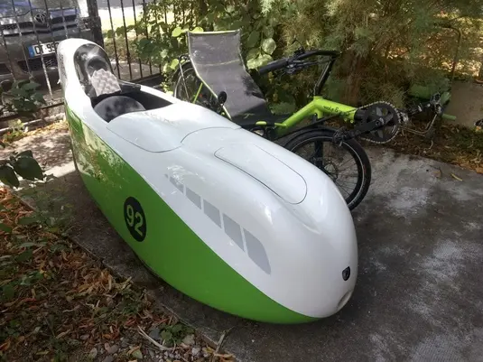 A green and white BÜLK velomobile with the number 92 on its side, parked outdoors with its top hatch open. The aerodynamic teardrop-shaped vehicle reveals its interior seating. A recumbent bicycle is visible in the background, and the setting appears to be a paved area with trees and a fence nearby.