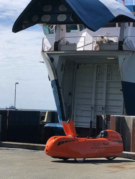 A bright orange velomobile with a sleek, aerodynamic design exits a ferry onto a dock. The ferry’s large open bow door is raised, revealing the interior ramp. The velomobile has a black canopy, small wheels, and is branded with the name 