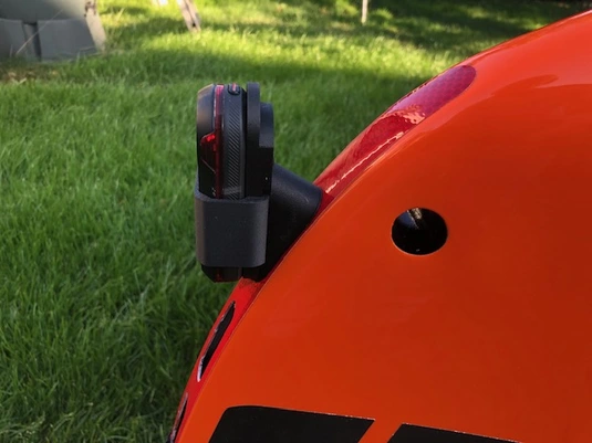 Detail shot of an accessory mounted on the orange velomobile's exterior. A small black device, possibly a light, camera, or sensor, is attached to the side of the bright orange shell. A circular ventilation hole is visible in the vehicle's body. The background shows green grass in a yard setting. This appears to be specialized equipment for the human-powered vehicle.