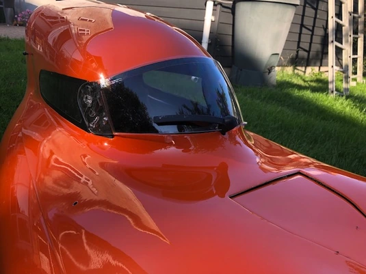 Close-up view of the front section of a bright orange velomobile featuring a sleek aerodynamic design with a black tinted windshield/visor area. The highly polished shell has a glossy finish that reflects sunlight. The vehicle sits on grass with household items visible in the background. The streamlined front showcases the vehicle's smooth contours and racing-inspired design.