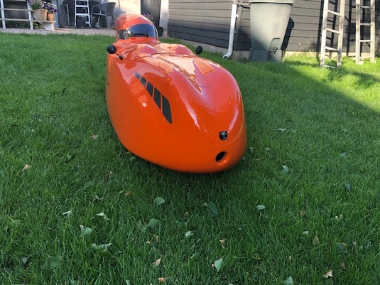 A bright orange velomobile with a sleek, aerodynamic design sits on a green lawn. The craft has a smooth, glossy finish with small vents or openings on its surface, possibly for ventilation or visibility. In the background, there's a house with beige siding, a ladder leaning against it, and what appears to be a trash bin. The setting appears to be someone's backyard or garden area.