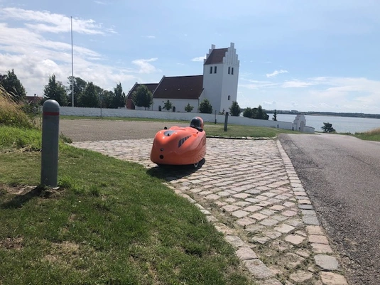 A bright orange velomobile with a sleek, aerodynamic design sits on a green lawn. The craft has a smooth, glossy finish with small vents or openings on its surface, possibly for ventilation or visibility. In the background, there's a house with beige siding, a ladder leaning against it, and what appears to be a trash bin. The setting appears to be someone's backyard or garden area.
