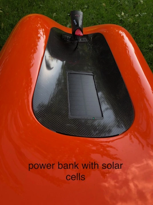A close-up of the rear section of an orange velomobile shows a carbon fiber surface with an integrated power bank featuring solar cells. A small green LED indicator light is visible on the solar panel. Above the panel, a rearview mirror with a red reflector is mounted. The text 