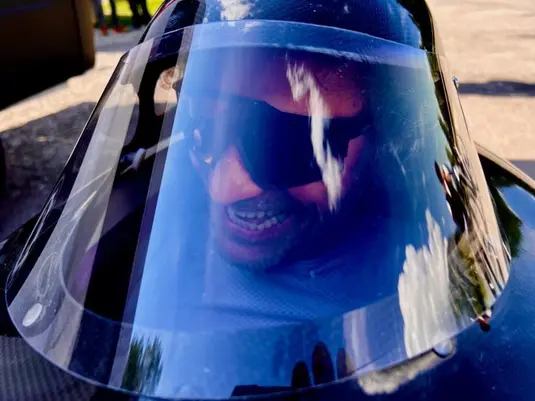 Close-up view through the transparent canopy of a velomobile showing a smiling person wearing sunglasses inside the cockpit. The clear windshield creates a slight reflection effect while protecting the rider inside the aerodynamic shell.
