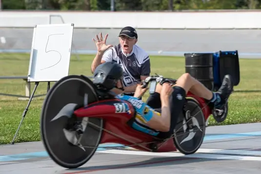 Recumbent bicycle racer in red racing position on a velodrome track while a coach enthusiastically gives instructions from trackside near a lap marker showing '5'