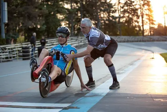 Coach assisting a recumbent bicycle racer at sunset on a velodrome track, demonstrating proper racing technique with golden light filtering through trees in the background.