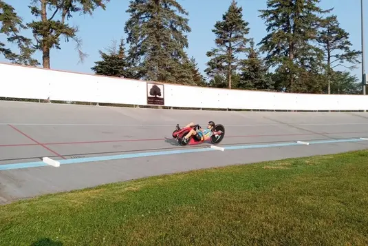 Aerial view of a recumbent bicycle racer leaning into a curve on a velodrome track, demonstrating the aerodynamic racing position against the white track walls and evergreen trees.